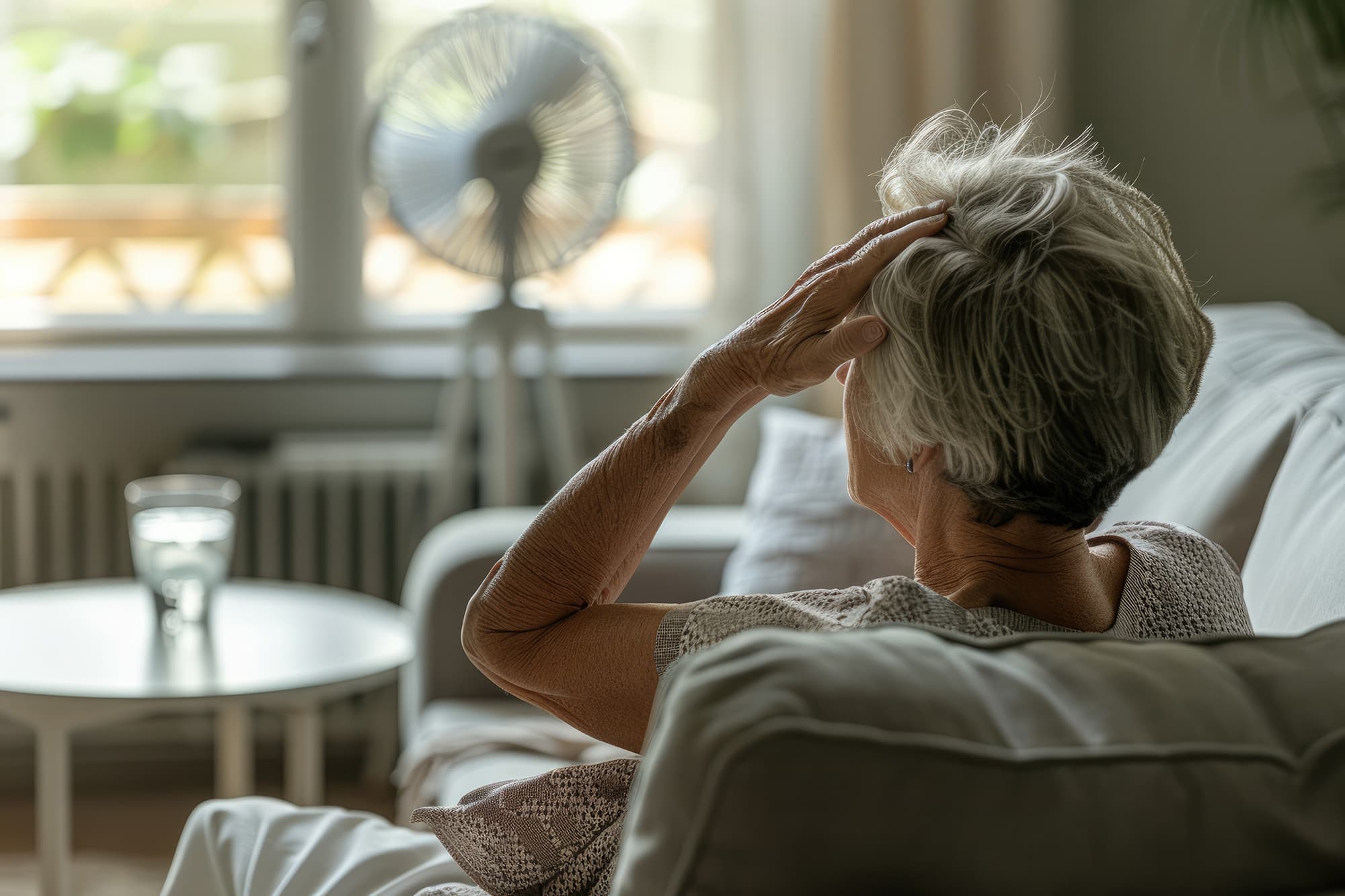 Äldre person i soffa bakifrån med fläkt håller sig för huvudet överhettad värmestress