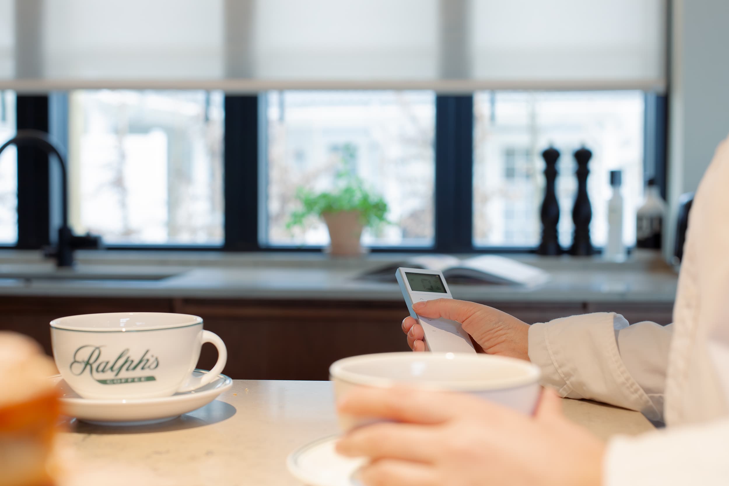 Person som håller en fjärrkontroll för solskydd i ett kök, med två kaffekoppar på köksö och ett fönster med rullgardin i bakgrunden